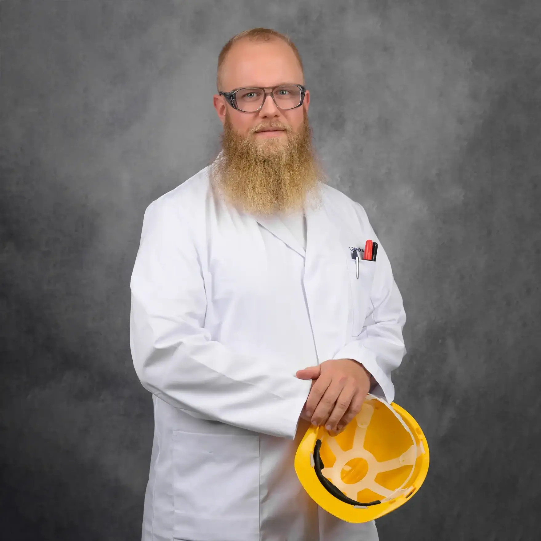 Ein Mann mit kurzen, hellen Haaren und einem langen Bart, der eine Sicherheitsbrille trägt in einem weißen Kittel und einem Sicherheitshelm in der Hand