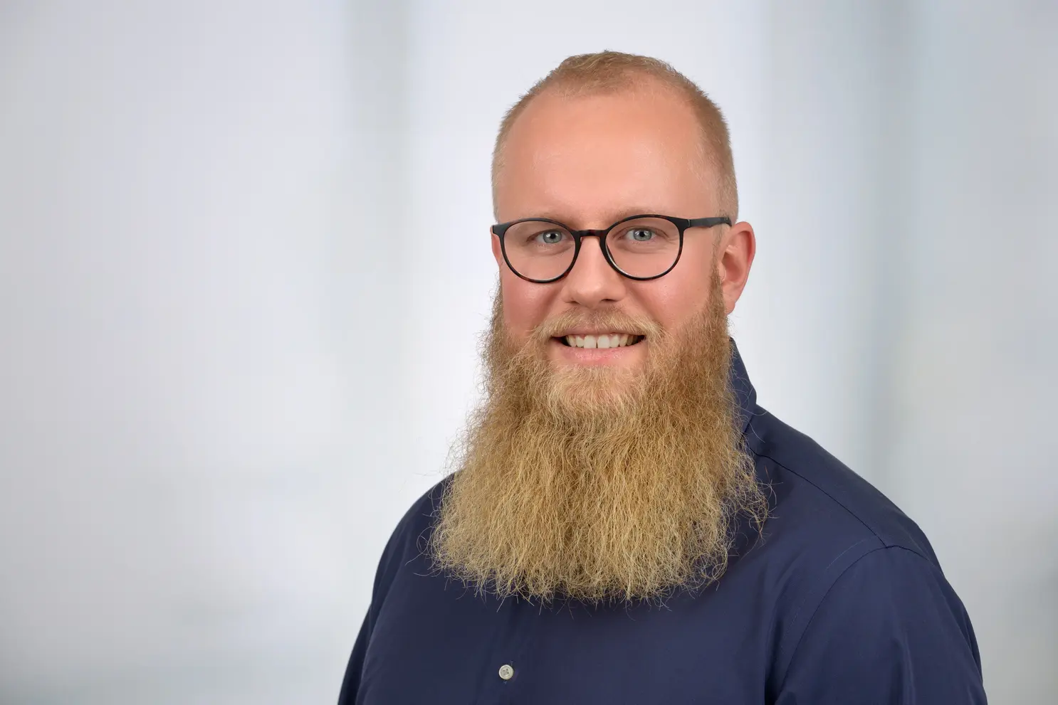 Ein Mann mit kurzen, hellen Haaren und einem langen Bart und einer Brille lächelt in die Kamera. Er trägt ein dunkelblaues Hemd