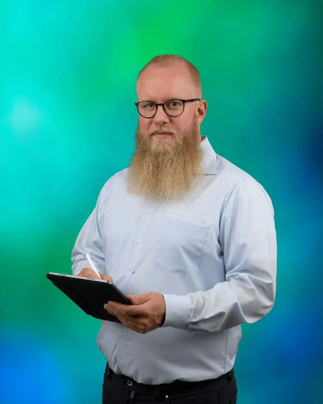 Ein Mann mit kurzen, hellen Haaren und einem langen Bart, der eine Brille trägt und ein Tablet in den Händen hält. Er trägt ein hellblaues Hemd und eine dunkelgraue Jeans.